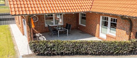 A brick house with a tiled roof, a patio with a table and chairs, and a hedge in front.
