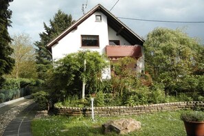 Ein eingeschossiges Haus mit geneigtem Dach, umgeben von einem Garten mit verschiedenen Pflanzen und einer Steinmauer.
