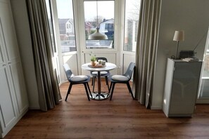 A dining area with a round table and four chairs, a small side table with a plant, and a refrigerator with a lamp on top. - Holiday apartment with 1 bedroom (Cuxhaven)