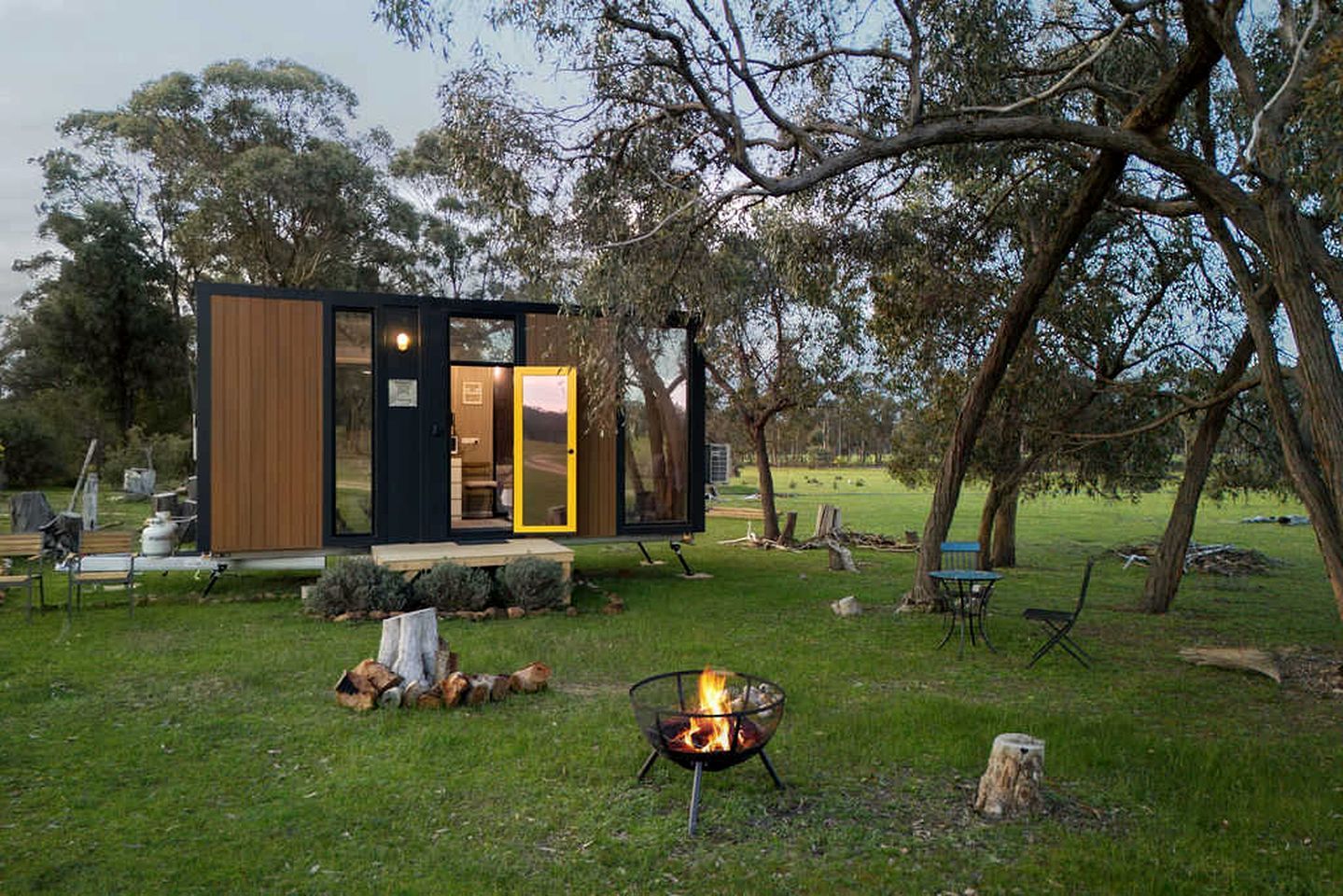 Een moderne cabin met een glazen deuropening, een vuurkuil en een gazon met bomen.