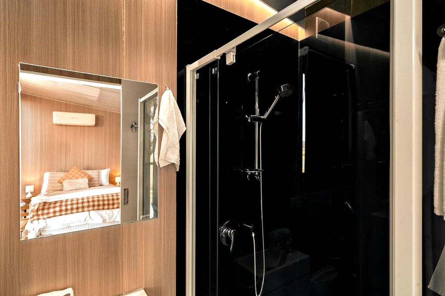 A hotel room with a large mirror reflecting a bed, a shower with a glass door, and a towel hanging on the door.