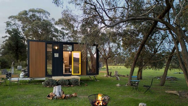 Uma cabana moderna com entrada por porta de vidro, lareira e gramado com árvores.