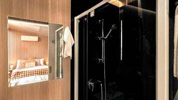 A hotel room with a large mirror reflecting a bed, a shower with a glass door, and a towel hanging on the door.