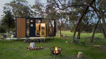 A modern cabin with a glass door entrance, a fire pit, and a grassy yard with trees.