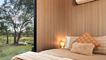Een slaapkamer met een groot raam, een bed met een zacht kussen, een nachtkastje met een lamp, en uitzicht op een gazon met bomen.