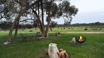 An outdoor seating area with chairs and a table, a fire pit, and a grassy field with trees in the background.