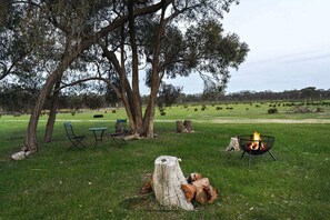 An outdoor seating area with chairs and a table, a fire pit, and a grassy field with trees in the background.