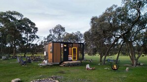 A small wooden cabin with a yellow door, surrounded by a grassy area with trees and a fire pit.