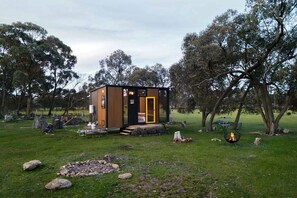 Una cabaña de madera pequeña con puerta amarilla, rodeada de césped, árboles y un hogar.