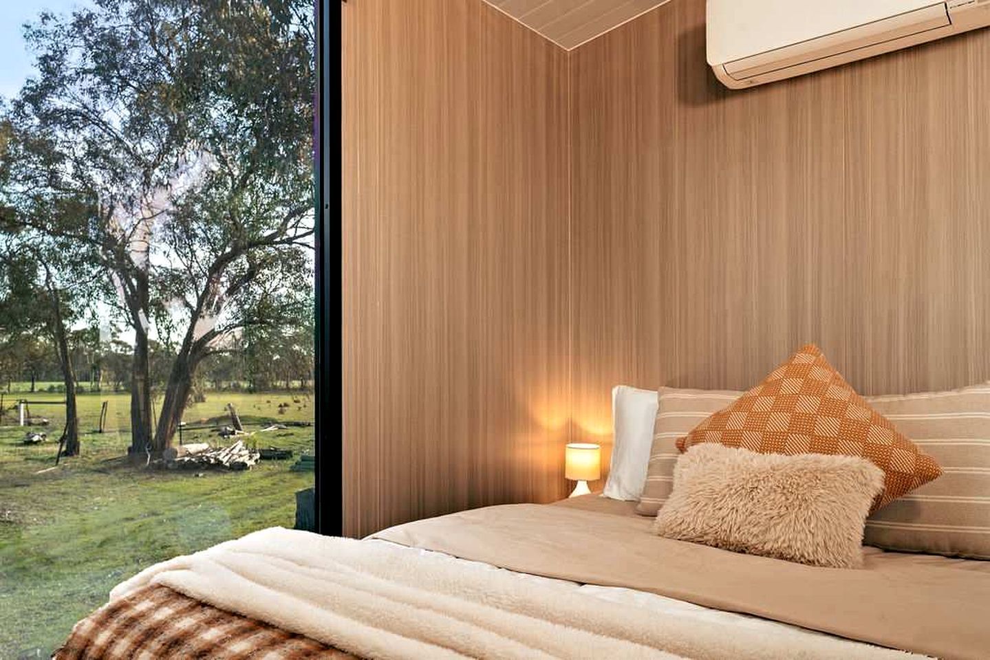 Une chambre avec une grande fenêtre, un lit doté d’un oreiller moelleux, une table de chevet avec une lampe, et une vue sur un espace verdoyant avec des arbres.