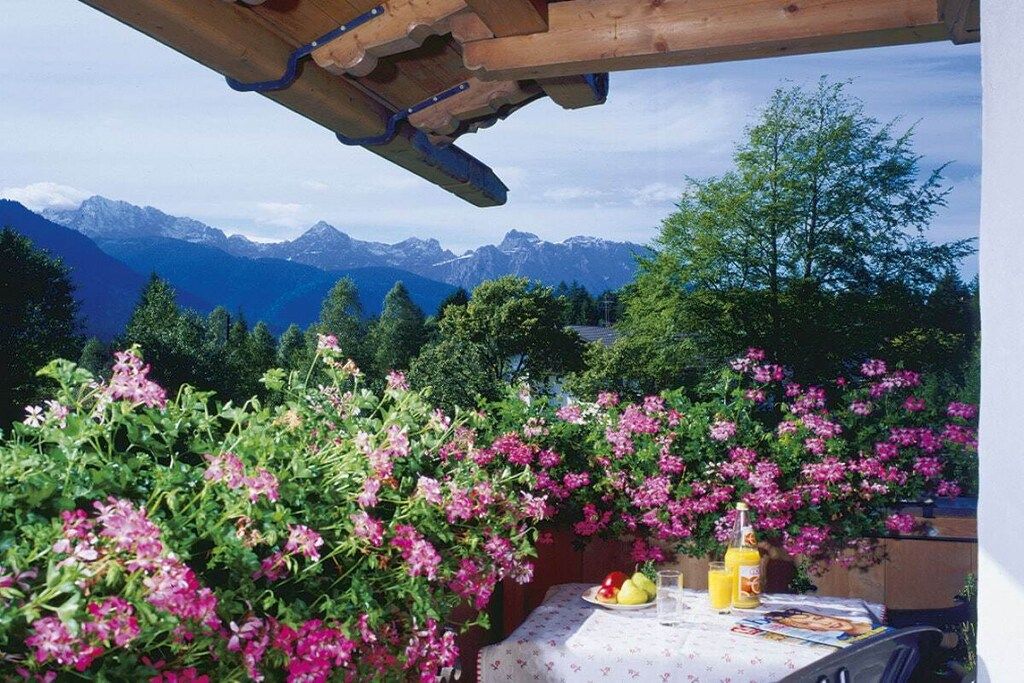 A table with a tablecloth, a bottle, and a glass, set against a backdrop of mountains and flowering bushes.