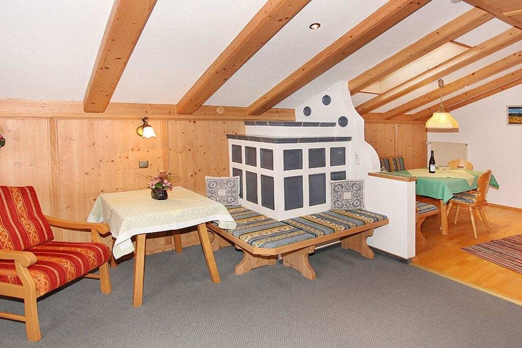 A cozy room with a wooden ceiling, a bench with patterned cushions, a small table with a vase, and a dining area with chairs and a table.
