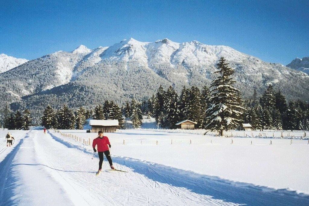 Schneebedeckte Landschaft mit einer Person beim Skifahren, einem Berg im Hintergrund und einigen Gebäuden.