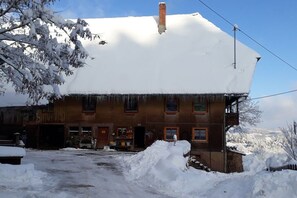 Ein zweistöckiges Gebäude mit verschneitem Dach und Schornstein. Im Erdgeschoss ist ein Eingang mit Tür und Fenstern zu sehen.