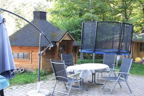 Eine Terrasse im Freien mit Tisch und Stühlen, einem Trampolin und einer Holzhütte im Hintergrund.