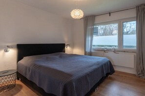 A bedroom with a bed, bedside lamps, a round side table, a window with curtains, and a hanging light fixture. - Holiday apartment Deichblick (Dangast)
