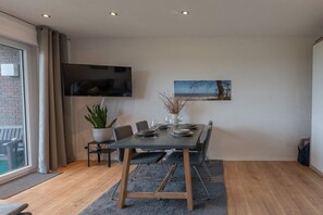 A modern dining area with a table set for four, a flat-screen TV mounted on the wall, and a large window with curtains. - Holiday apartment Deichblick (Dangast)