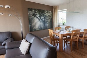 Ein modernes Wohnzimmer mit Ledersofa, Esstisch aus Holz und Stühlen. Ein großes Gemälde zeigt eine Waldszene.