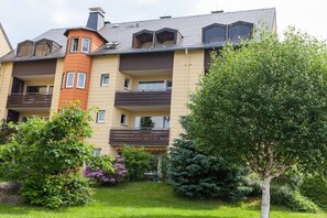 Ein mehrstöckiges Wohngebäude mit Balkonen, ein Gartenbereich mit verschiedenen Sträuchern und Bäumen und einem wolkenlosen Himmel.
