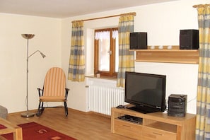 A room with a television on a wooden stand, a chair, a radiator, and curtains. - Holiday apartment with 2 bedrooms (Altenau)