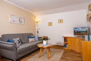 A living room with a grey sofa, a small round wooden table with a vase of flowers, a bookshelf, and a television on a wooden cabinet. - Holiday apartment with 1 bedroom (Bad Hindelang)
