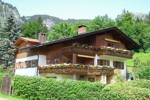 A two-story chalet with a balcony adorned with flowers, set against a mountainous backdrop. - Holiday apartment with 1 bedroom (Bad Hindelang)
