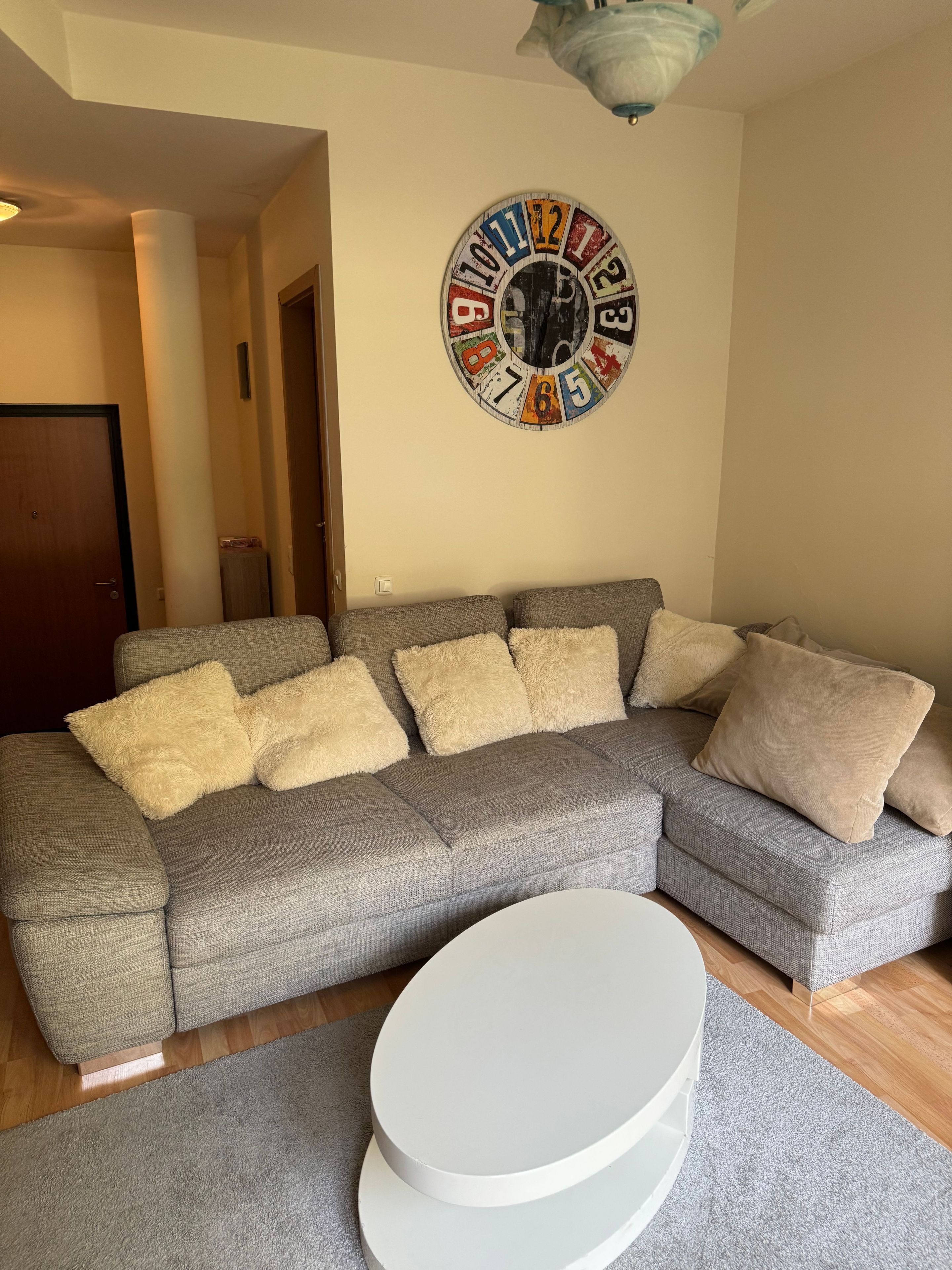 Een woonkamer met een grijze bank, beige en bruine kussens, een witte salontafel en een wandklok.