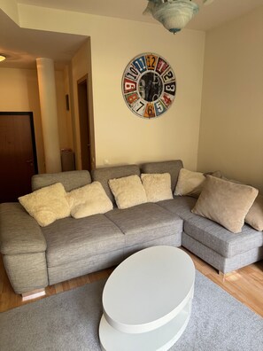 Una sala de estar con un sofá gris, cojines color beige y marrón, una mesa de centro blanca y un reloj de pared.
