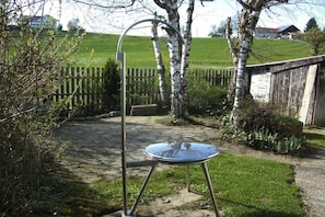 Een metalen tafel met een glazen blad in een tuin met een geplaveid pad, omringd door begroeiing en een houten hek.