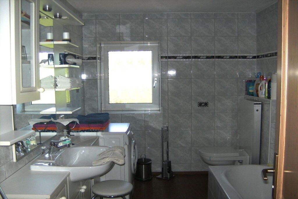 Een badkamer met een wastafel, toilet en bad. Er is een raam boven de wastafel en planken met toiletartikelen.
