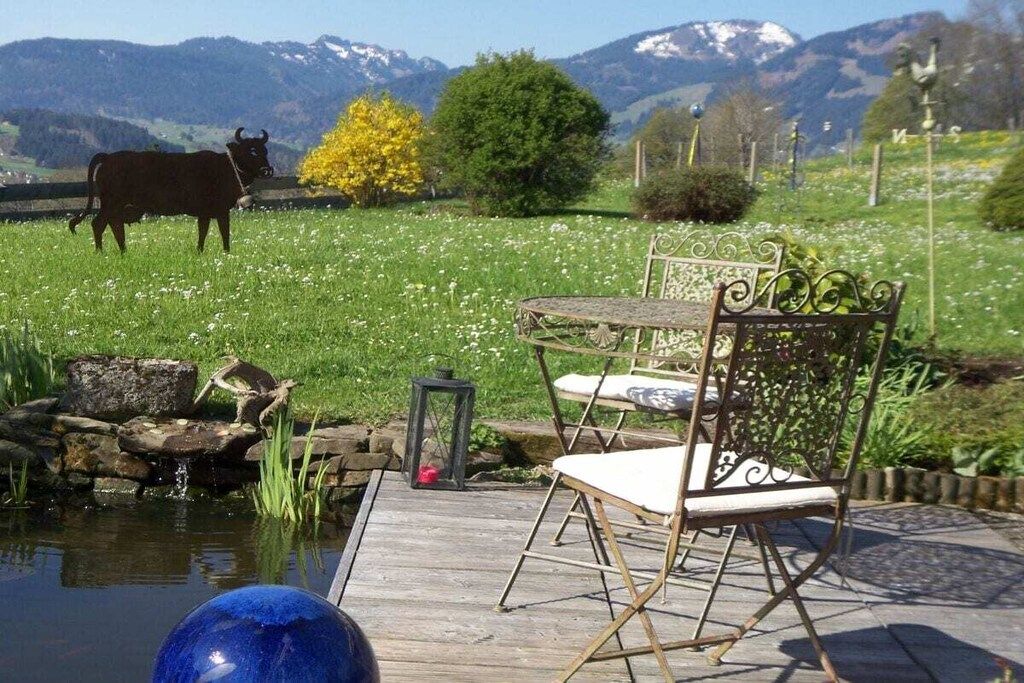 A rustic outdoor seating area with a wooden table and metal chairs, a small pond, and a cow in a field with mountains in the background.