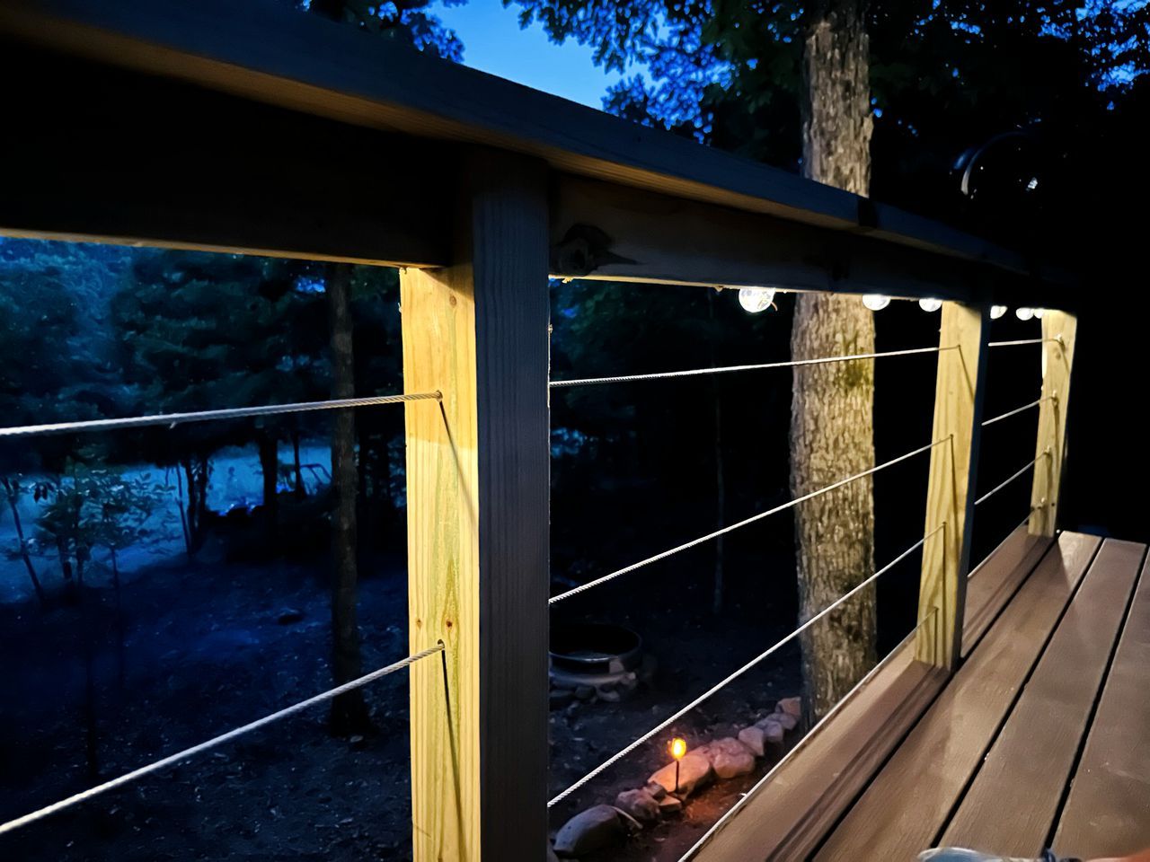 A wooden deck with a railing, surrounded by trees and a campfire.