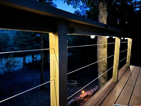A wooden deck with a railing, surrounded by trees and a campfire.