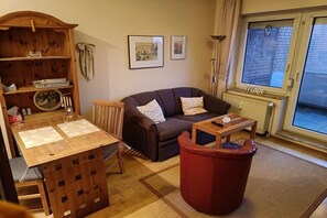 Un salon chaleureux avec une table en bois, un canapé, un pouf rouge et une fenêtre donnant sur un immeuble en briques.