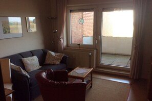 Ein Wohnzimmer mit einer Couch, einem Sessel und einem kleinen Tisch. Ein Fenster bietet Blick auf eine Ziegelwand, und eine Tür führt nach draußen.