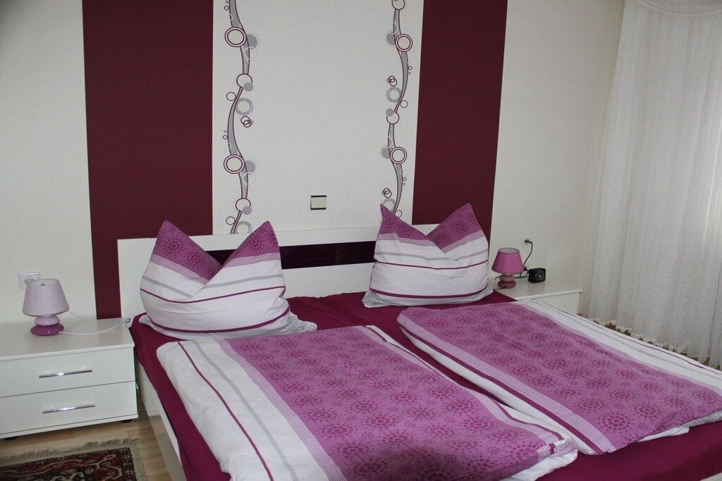 Un dormitorio con una cama, dos almohadas, una mesita de noche con una lámpara y un mueble auxiliar con una lámpara.