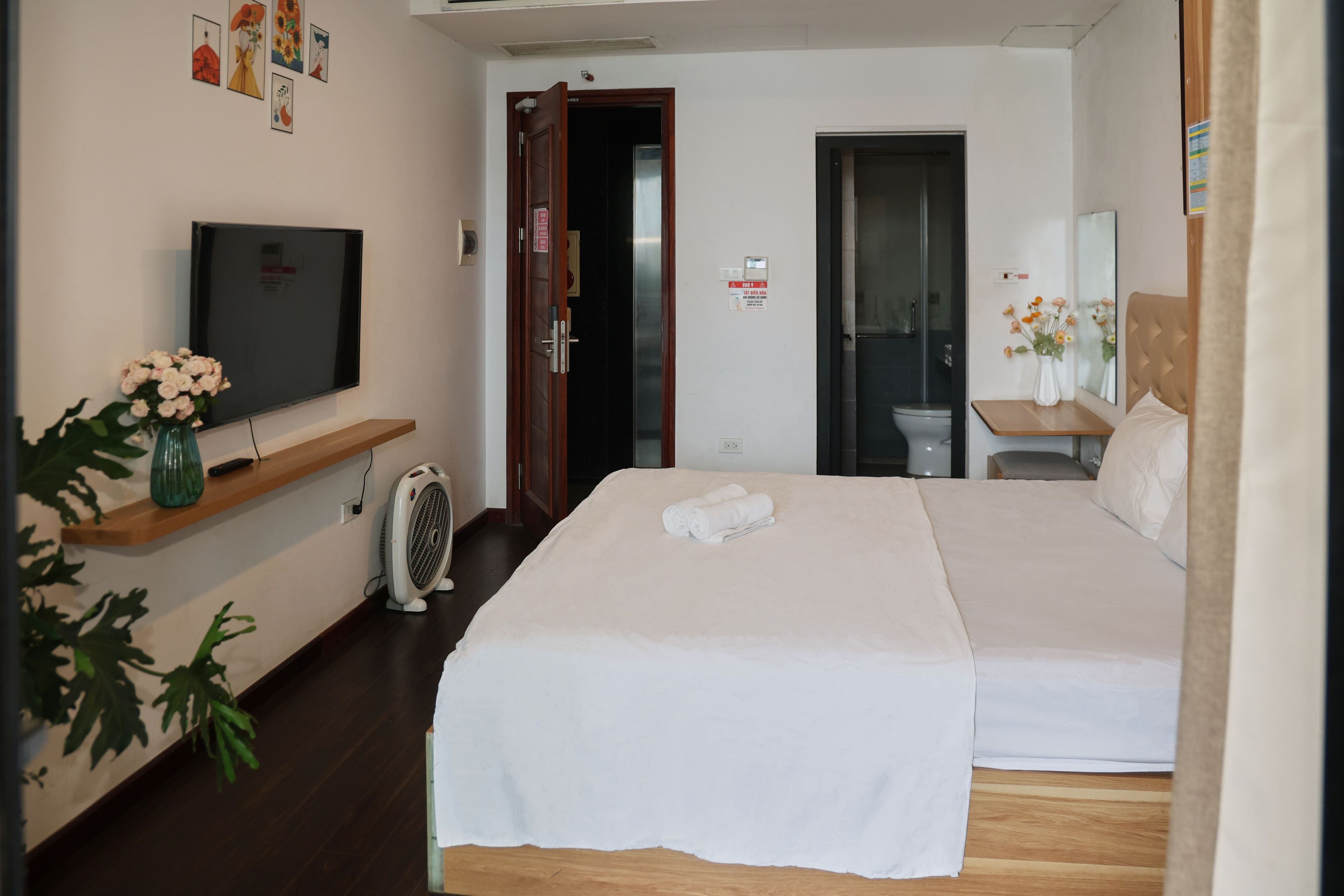 A hotel room with a bed, a TV, a fan, a bathroom, and a small table with a vase.