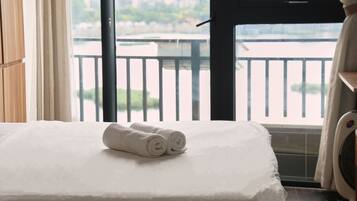 A hotel room with a bed, a window with a view of a body of water and greenery, and a balcony with a railing.