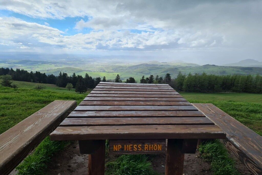 Una mesa de picnic de madera con un cartel que dice 'NP HESS RHÔN' en un campo cubierto de césped, con vistas a colinas onduladas y un cielo nublado.