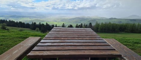 Una mesa de picnic de madera con un cartel que dice 'NP HESS RHÔN' en un campo cubierto de césped, con vistas a colinas onduladas y un cielo nublado.
