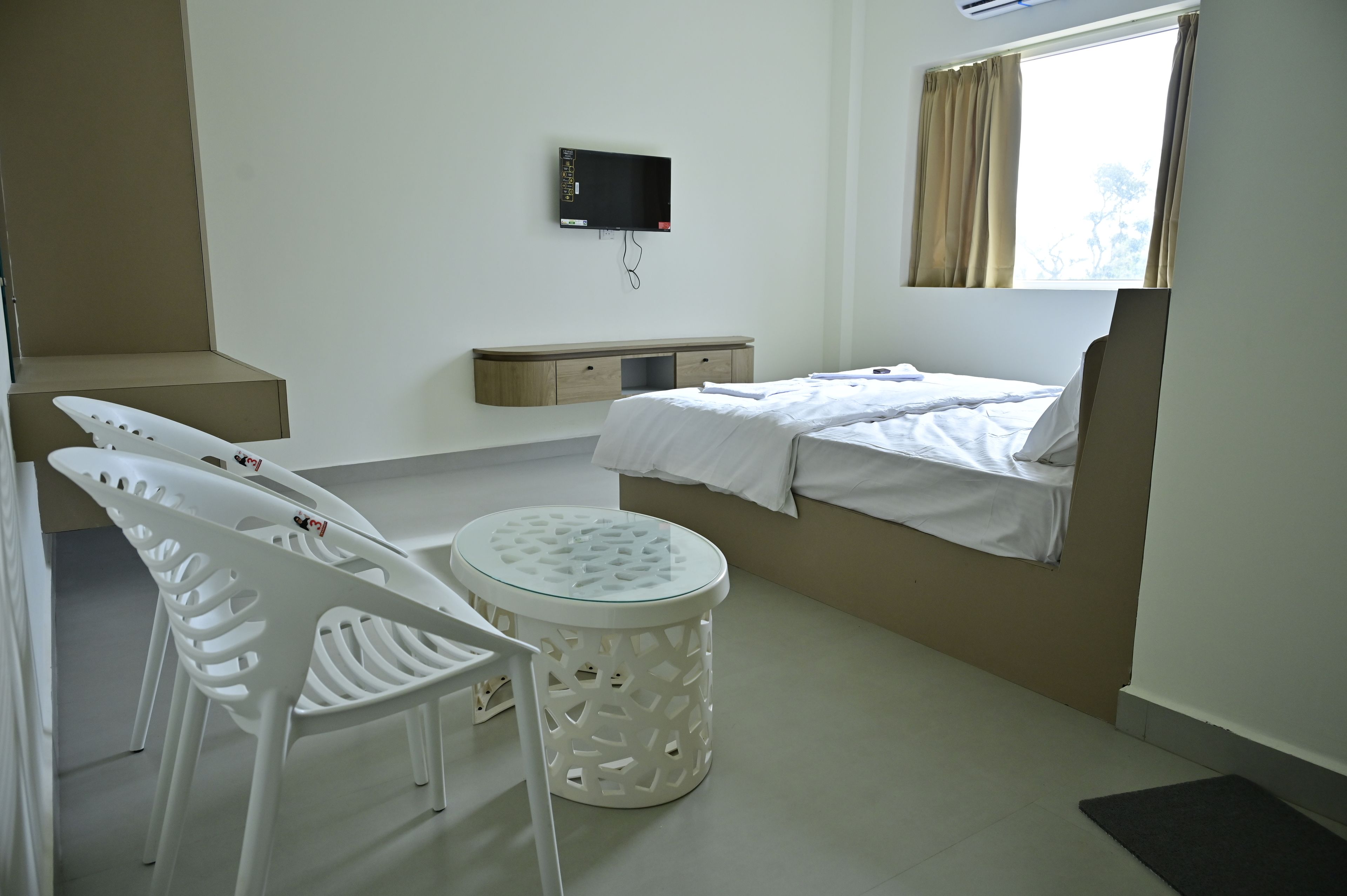 A hotel room with a bed, a small table, a chair, a TV mounted on the wall, and a window with curtains.