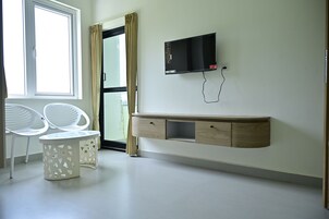 A room with a TV mounted on the wall, a wooden cabinet, two white chairs, and a window with curtains.