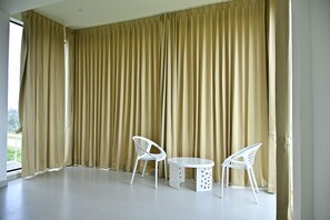 Una habitación con cortinas color beige, piso blanco y dos sillas blancas junto a una pequeña mesa.