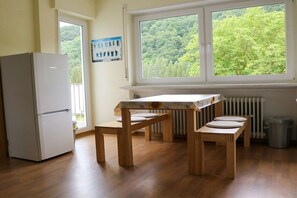 Eine Küche mit Kühlschrank, Holztisch und Bank, ein Fenster mit Blick auf Bäume und ein Heizkörper.