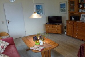 A living room with a sofa, a wooden table with a vase and a magazine, a television on a wooden cabinet, and a door. - Nolte - Guesthouse Holnis (Glücksburg)