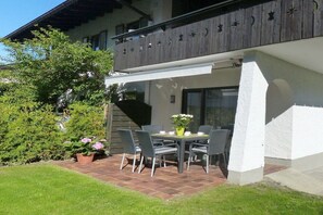 Een terras buiten met een tafel en stoelen, omringd door een tuin met een boom en plantenpotten.