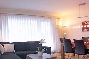 A living room with a sofa, a coffee table with a plant, a dining area with chairs and a table, and a large window with curtains. - Holiday apartment with 3 bedrooms (Garmisch-Partenkirchen)