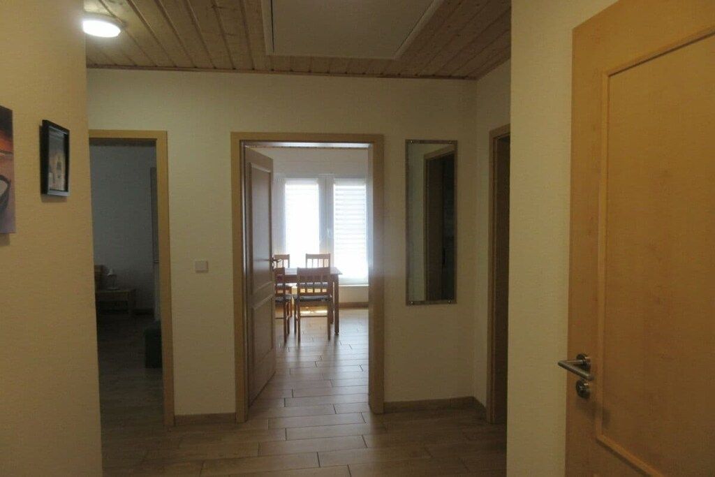 Een gang met een houten deur aan de rechterkant, een eetgedeelte met stoelen en een tafel in het midden, en een deur naar een andere kamer aan de linkerkant.