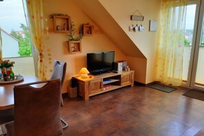 A living room with a television on a wooden stand, a dining table with chairs, and a window with curtains. - Kühlungsborn West Beach (Kühlungsborn)