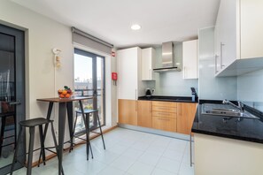 A modern kitchen with wooden cabinets, a black countertop, and a stainless steel sink. There is a small table with a bowl of fruit and two stools. - Privilege 2-Bedroom Apartment with Rooftop Pool Terrace, Village Marina - Olhão (Olhão)